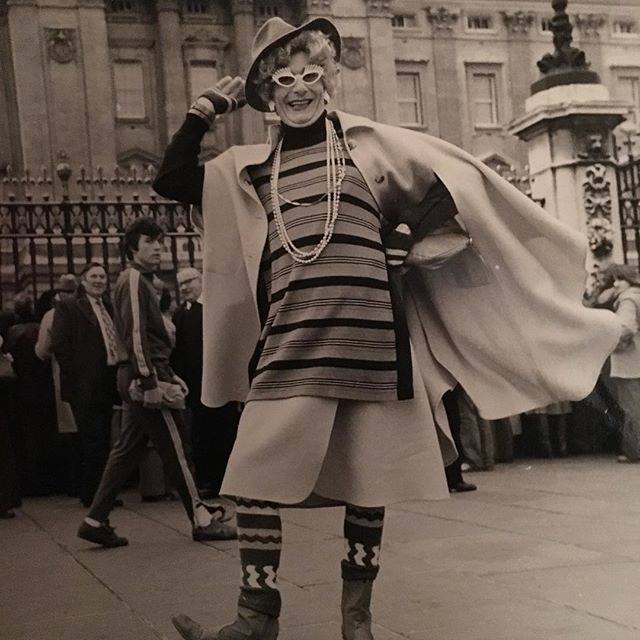 A black and white photo of an early version of Dame Edna. Humphries poses with one hand up wearing a hat