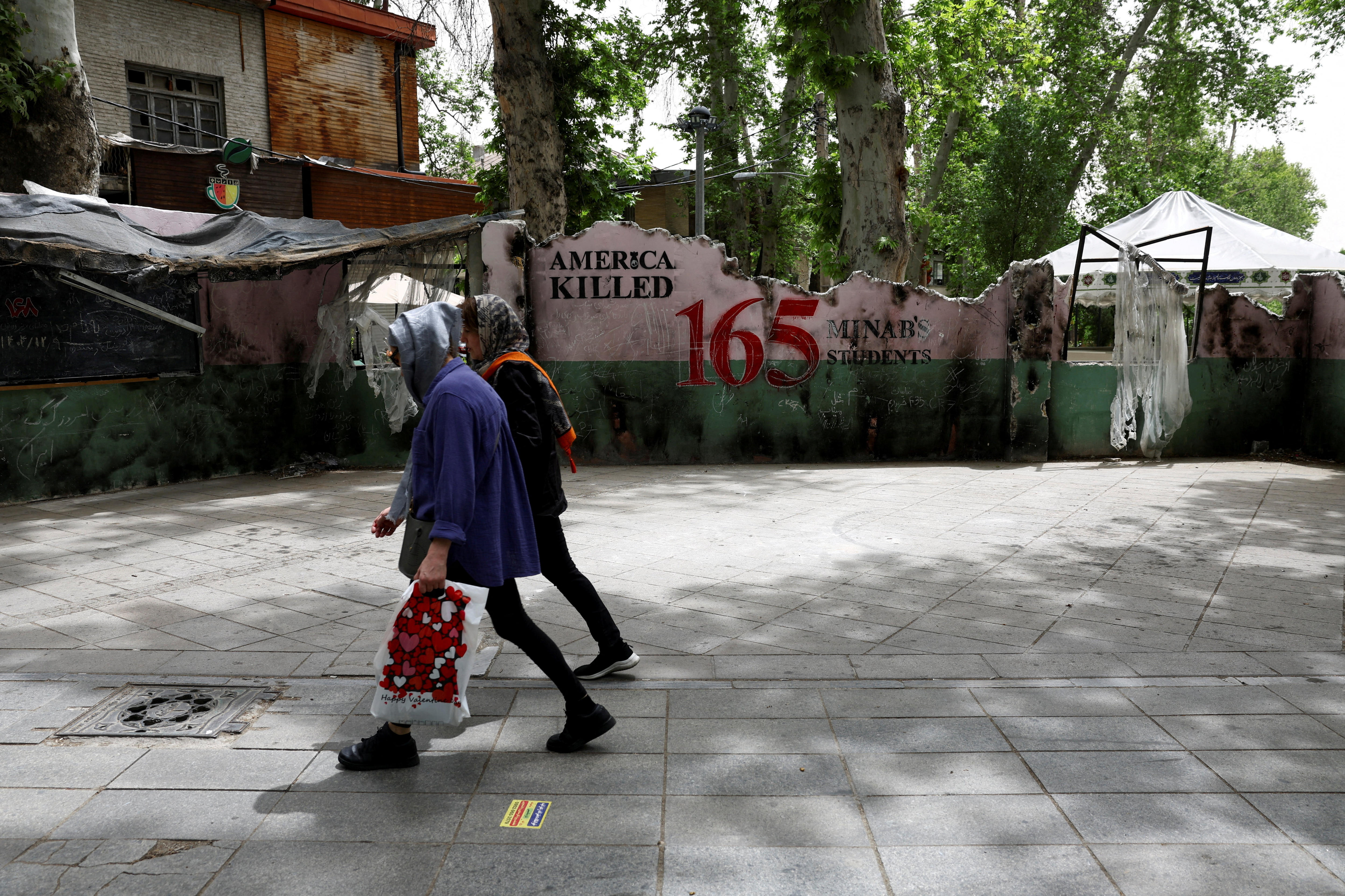 Two people walking past a wall that says "America killed 165 Minab's students".