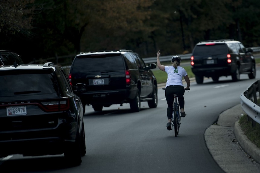 Woman cycling on the side of the road gestures to a trail of three black SUVs