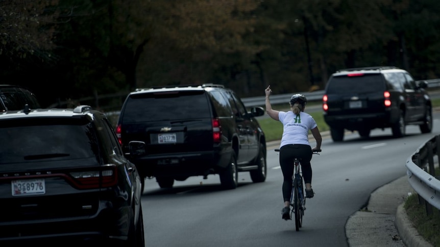 Woman cycling on the side of the road gestures to a trail of three black SUVs