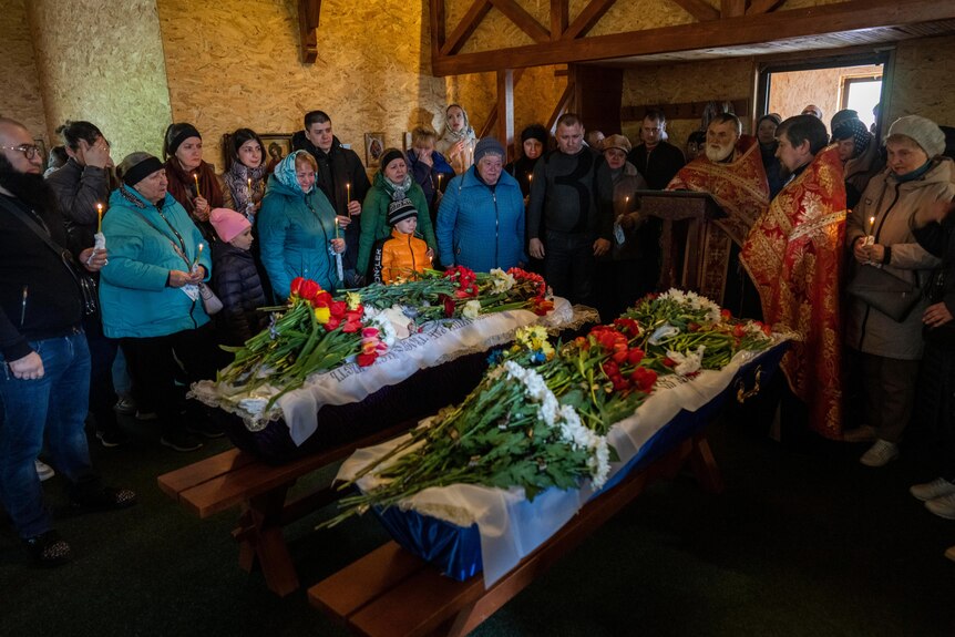 Adults and children stand around two coffins adorned with red, white and yellow flowers with long green stems.