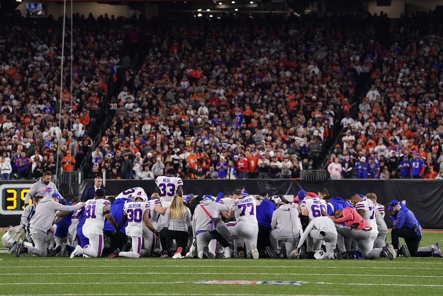 A group of Buffalo Bills NFL players kneel in a circle on the field with heads down during a game after a teammate's injury.