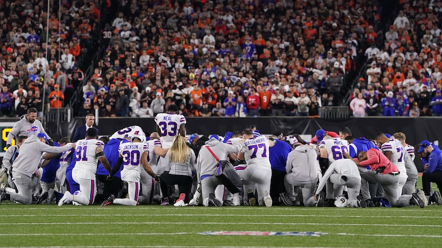 A group of Buffalo Bills NFL players kneel in a circle on the field with heads down during a game after a teammate's injury.