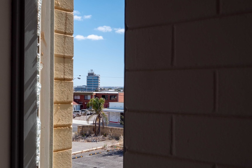 A  broken window reveals the view of a city, with a tall building in the distance under a blue sky.