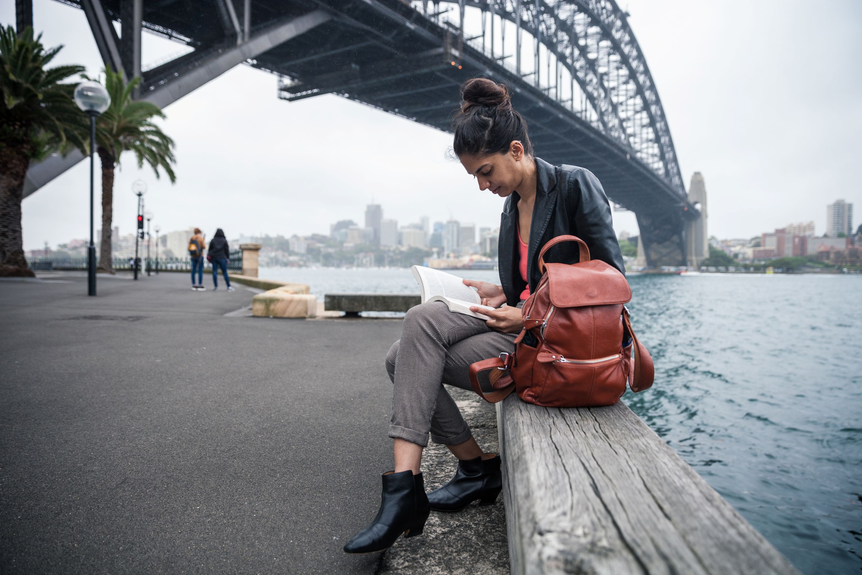 A woman reading a book as she sits on a timber bench, with the Sydney Harbour Bridge behind her.