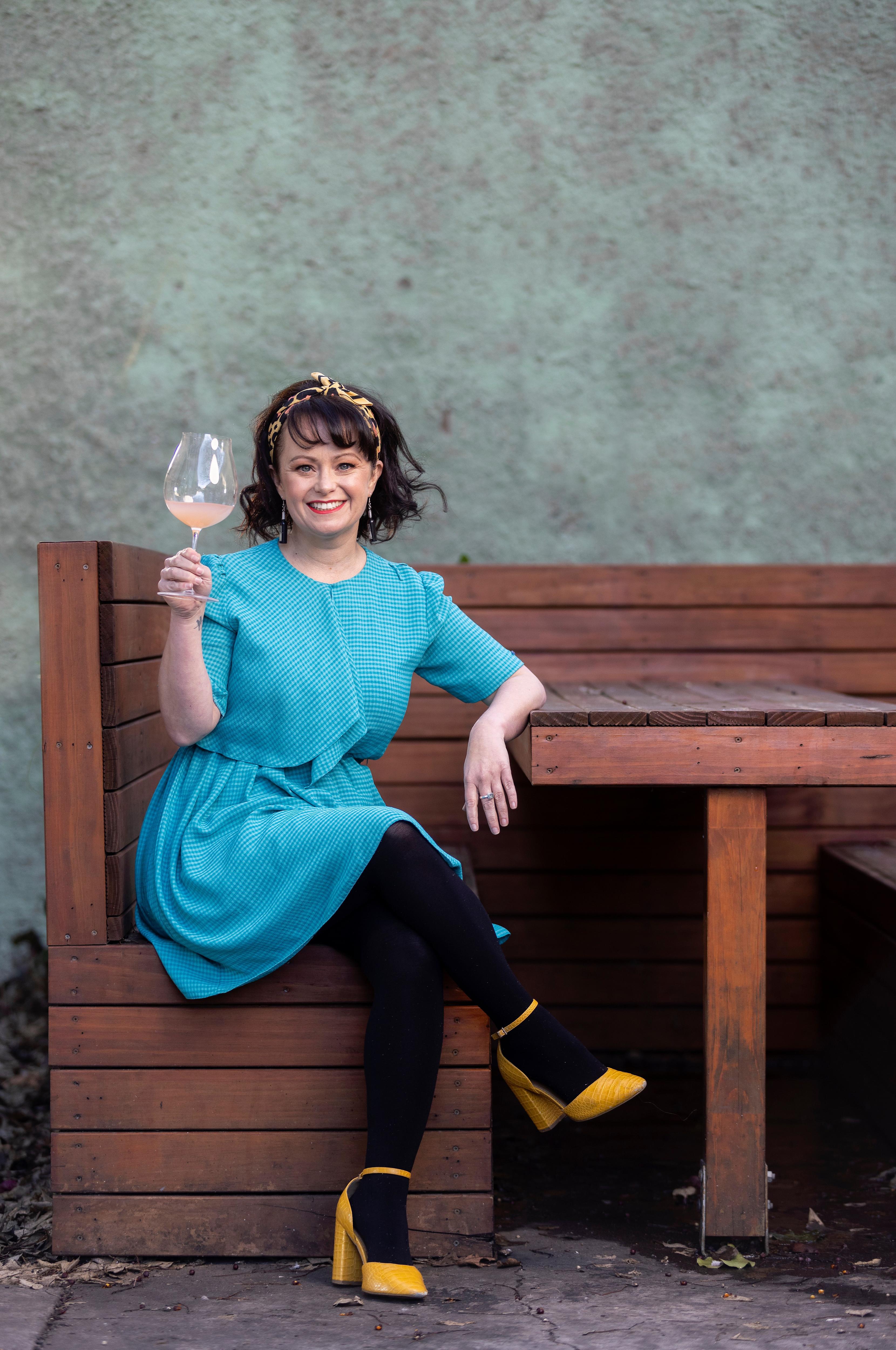Katie Spain sits at a table smiling at the camera holding a glass of wine. 
