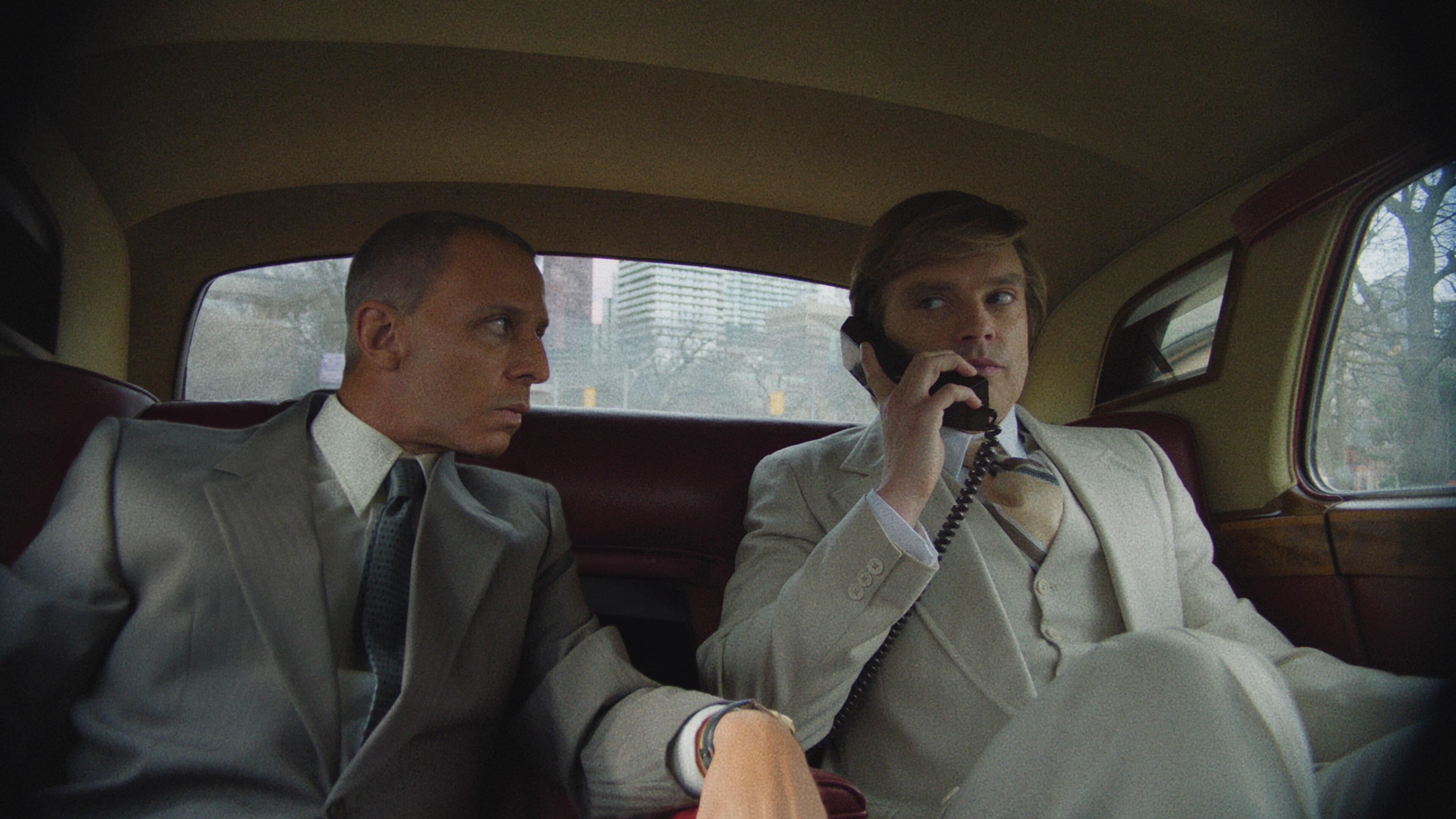Two men in grey suits sit in the back of a town car, one is on a landline phone
