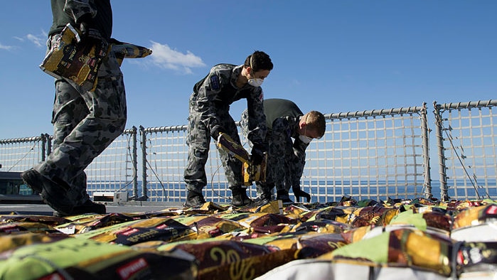 Dozens of small packets are lined up on the deck of a ship by men in navy uniforms and breathing masks.