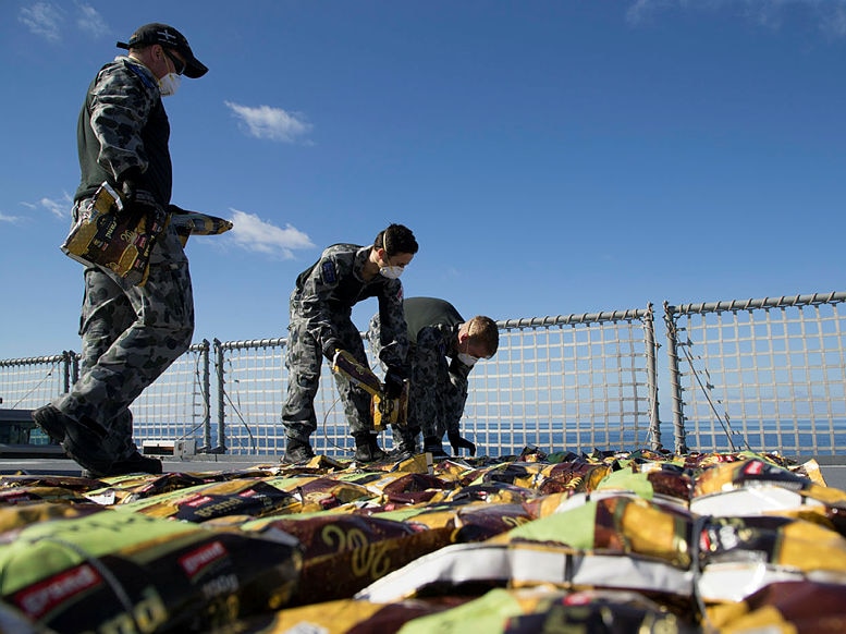 Dozens of small packets are lined up on the deck of a ship by men in navy uniforms and breathing masks.