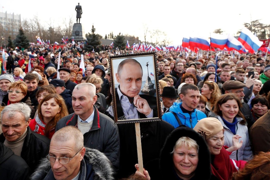 Thousands of people at a rally, one in foreground holds a framed photo of Putin