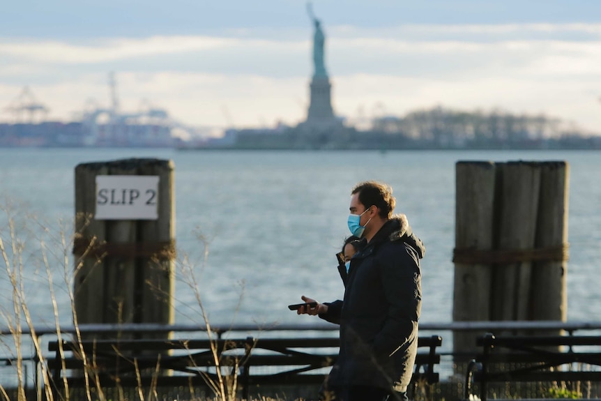 Two people wearing face masks walk with the Statue of Liberty in the background.