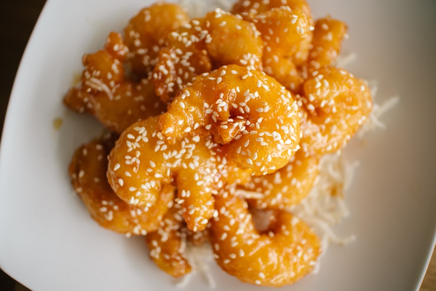 Golden battered and fried prawns are covered in white sesame seeds and stacked on a white plate.