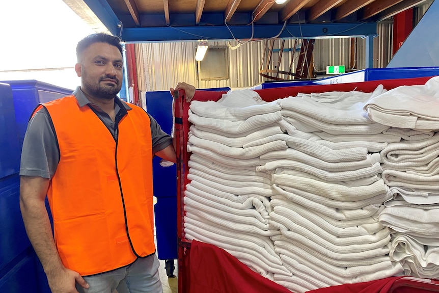 A man with short black hair wearing high visibility orange vest standing beside stack of white towels