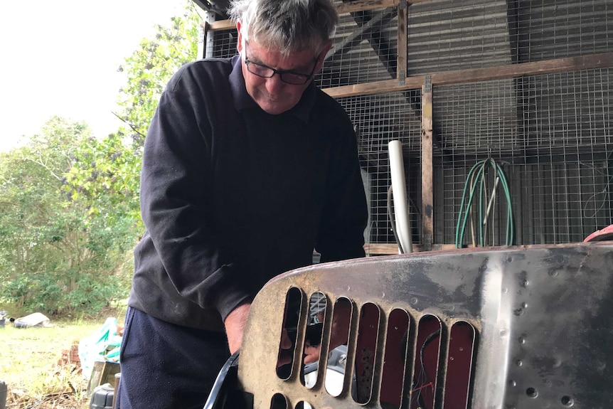 An elderly man holds a part of an old car he is restoring