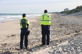 Two police officers with backs turned on the beach, with water on their left and a police car in the distance on their right.