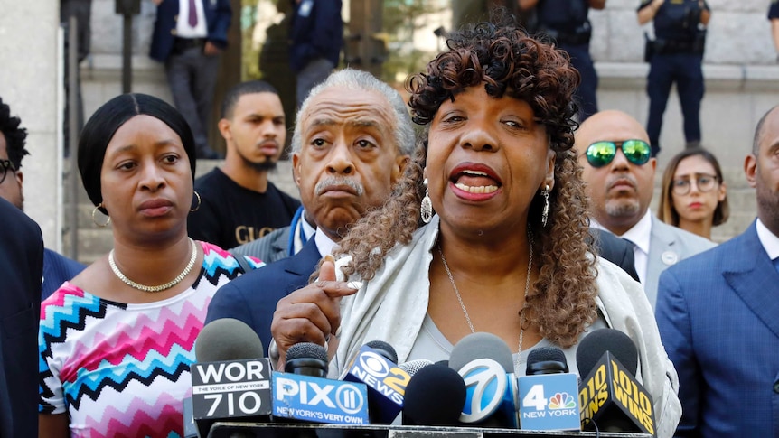 A crowd of African American people speak behind a raft of different coloured media microphones on the steps of a large office.