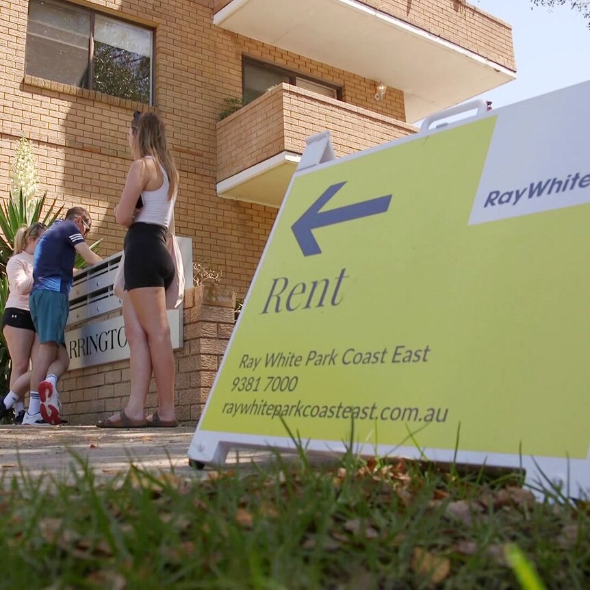 people standing outside an apartment block waiting to view it as a rent sign stands on the footpath