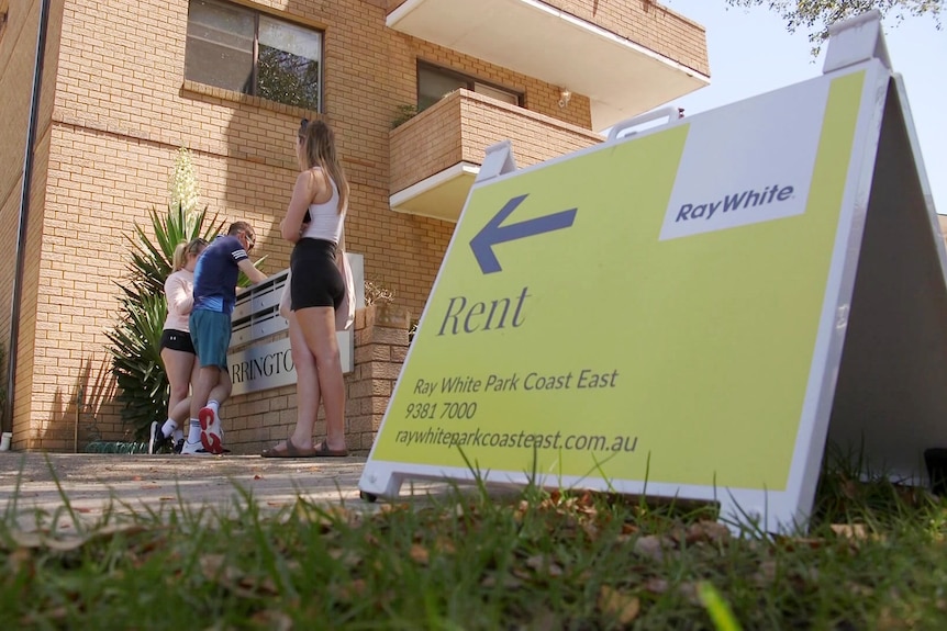 people standing outside an apartment block waiting to view it as a rent sign stands on the footpath