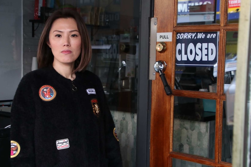 A woman stands at the door of her shop next to a closed sign.
