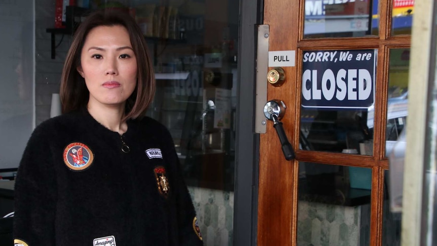 A woman stands at the door of her shop next to a closed sign.