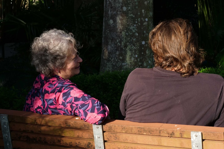 A mother and her adult son sit at park bench in afternoon sunlight.