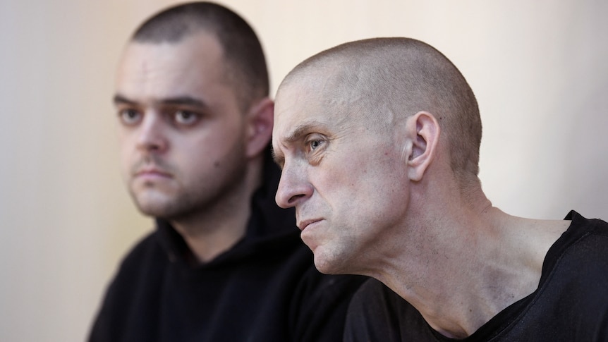 Two men sit in a sell and listen intently to someone out of frame. 