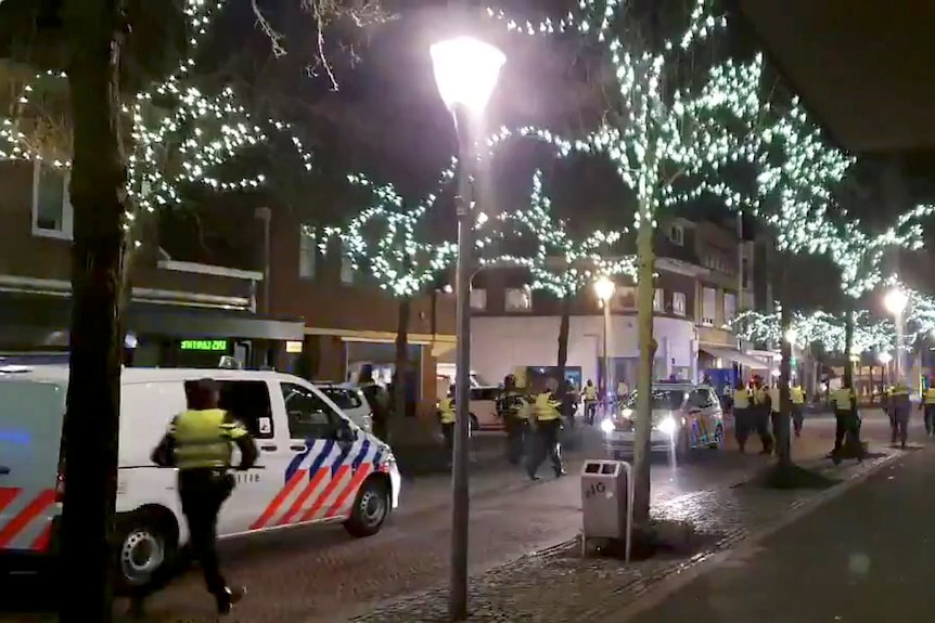 A police officer wearing yellow runs along a tree-lit city street towards colleagues and parked cars