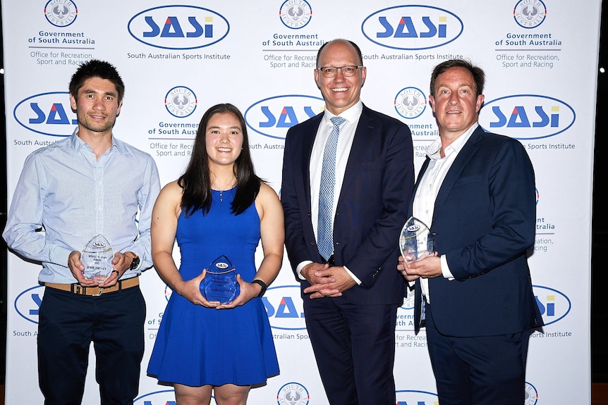Three men and a woman pose for a photo in front of a SASI banner, three of them holding glass award