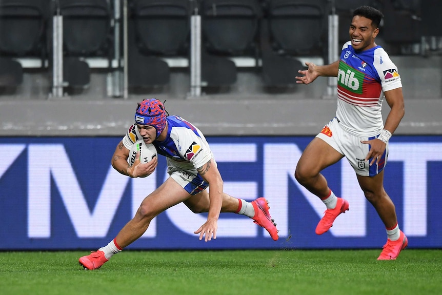 An NRL player dives over to score a try as his teammate grins in the background.