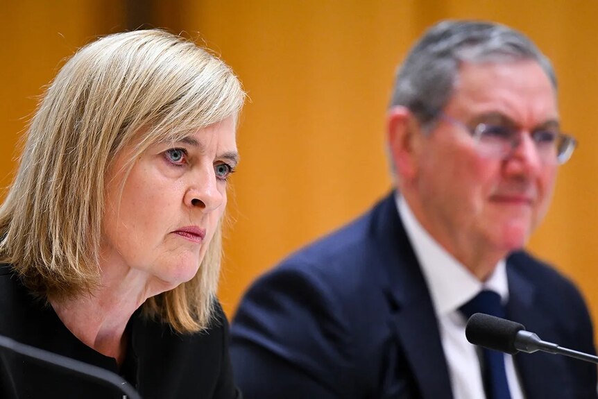 A blonde woman in black blazer with a serious expression sits at a microphone next to an older man in suit
