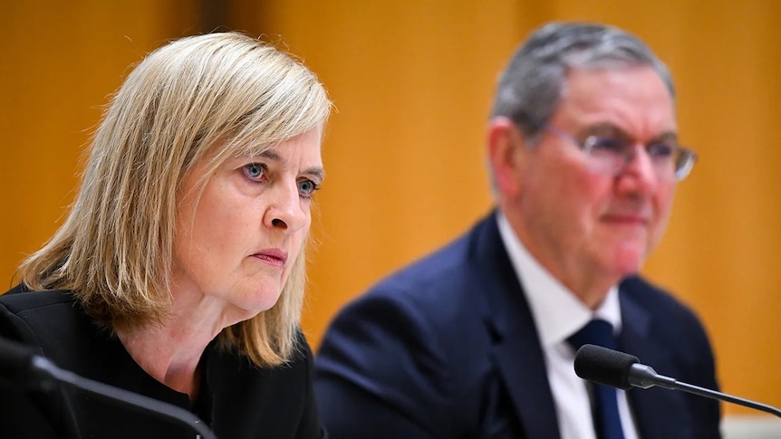 A blonde woman in black blazer with a serious expression sits at a microphone next to an older man in suit