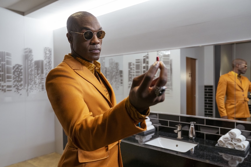 A 35-year-old man in glasses and yellow suit stands in a bathroom holding a red pill out