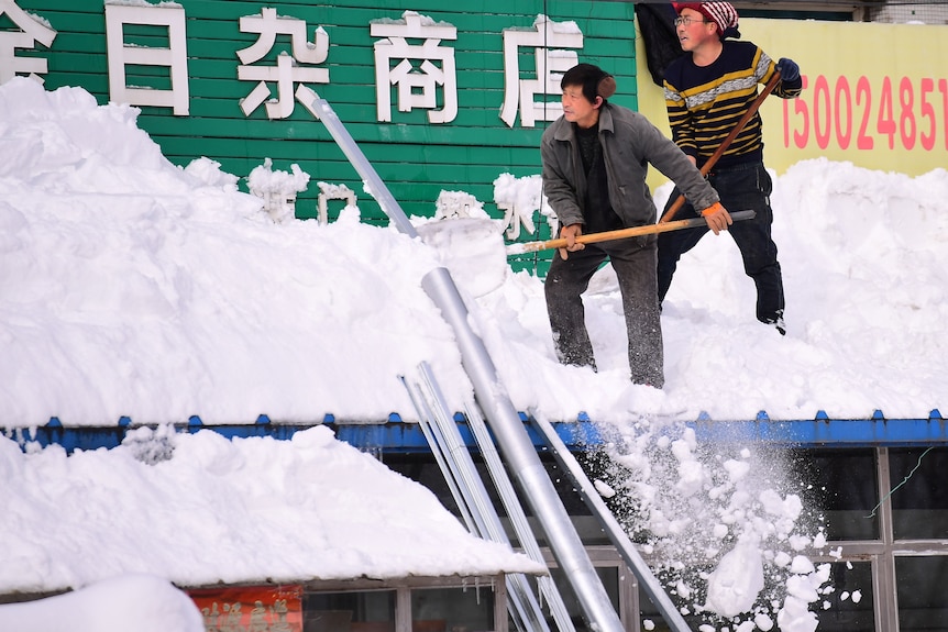 Two men shoveling snow off of a shop front roof