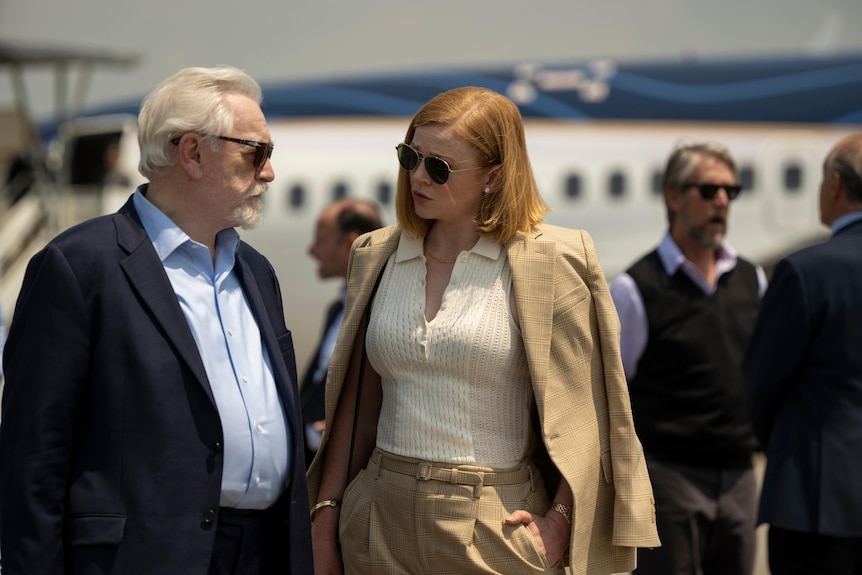 An older man and younger woman in conversation while wearing sunglasses.