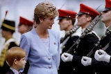 You view Princess Diana in a tailored, lavender blazer and matching top, walking alongside troops in red hats with Prince Harry.