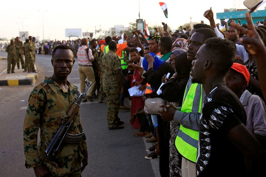 Hundreds of protesters stand in a line as the Sudanese military holds them back on a road.