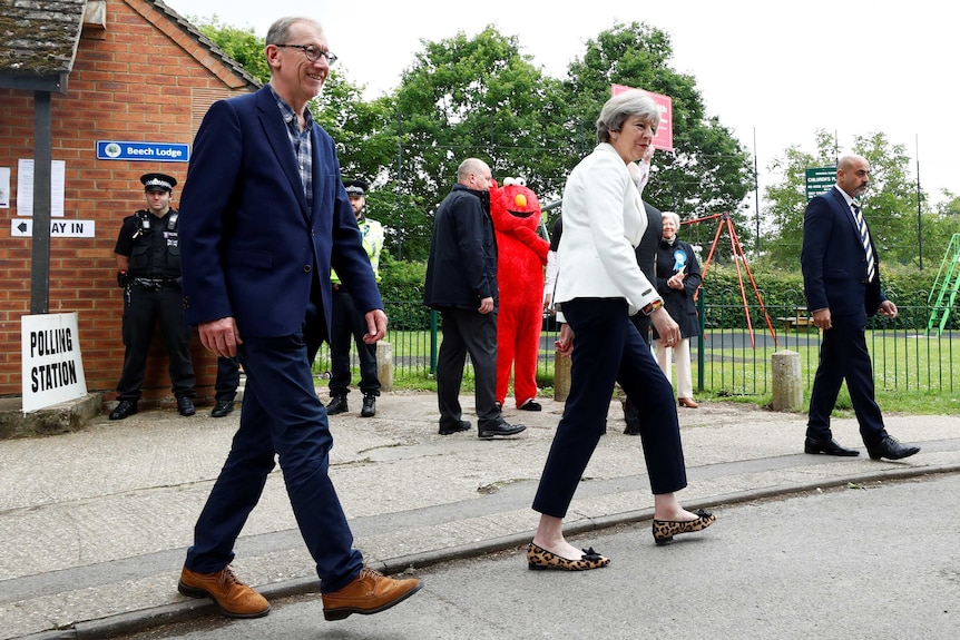 A woman wearing a white suit jacket and black pants walks past a man wearing a red Elmo suit.