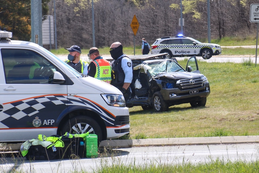 A vehicle is badly damaged, with its windscreen smashed, near a police van and police officers on the side of a street.