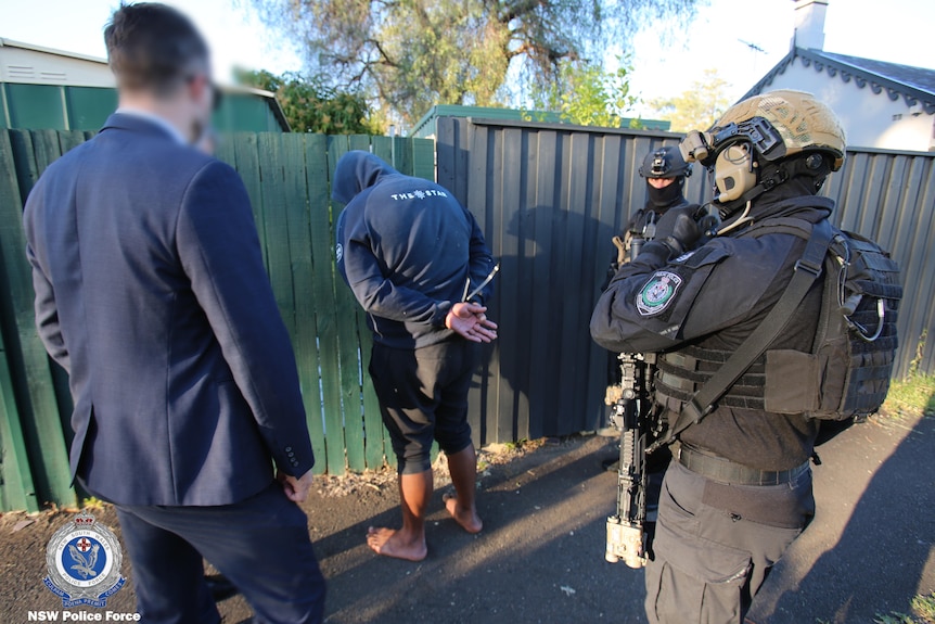 A man with a hoodie on faces a fence with his hands tied together and police around him