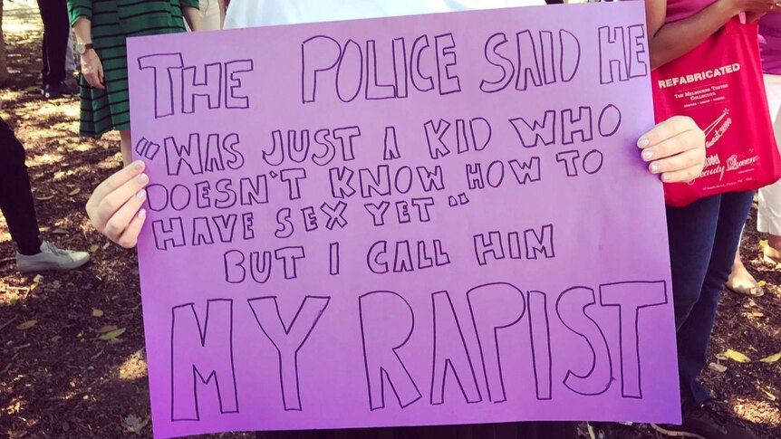 A woman holds a purple sign: "The police said he was just a kid who doesn't know how to have sex yet. But I call him my rapist."