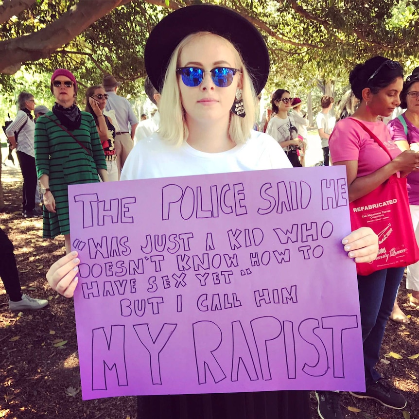 A woman holds a purple sign: "The police said he was just a kid who doesn't know how to have sex yet. But I call him my rapist."