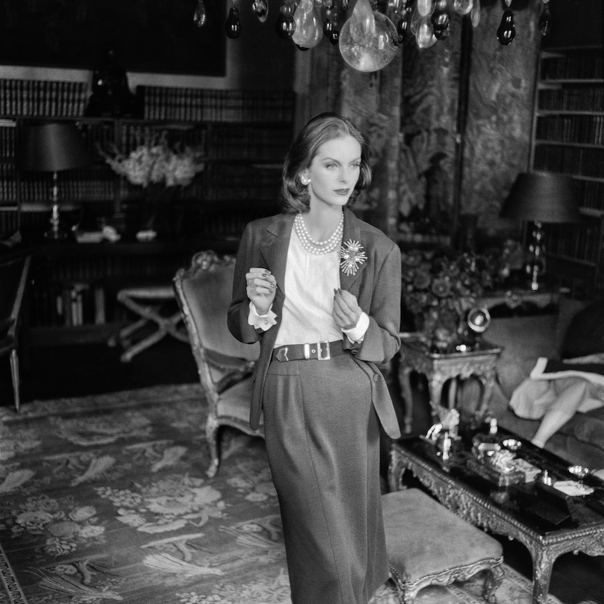 A black and white photo of a young woman in a Chanel suit in 1955, standing in a fancy room