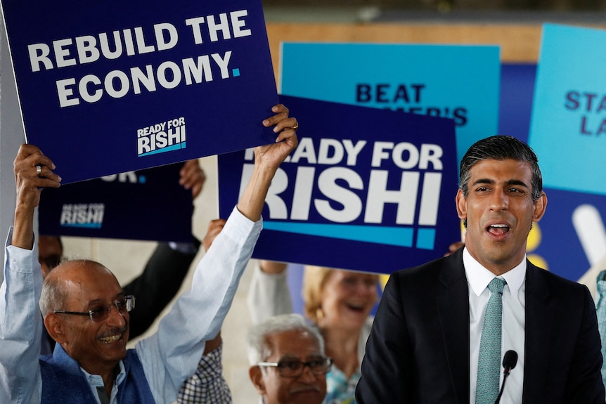 A man in a suit stands at a microphone and in the background people hold blue signs. 