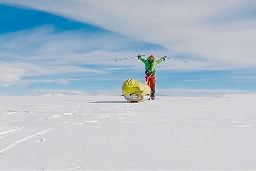 Colin O'Brady holds his ski poles in the air as he stands near his sled in the ice.