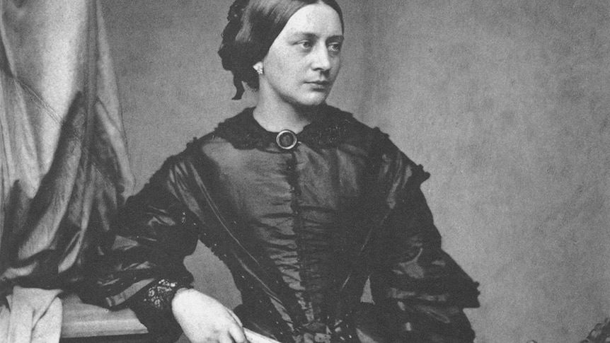 A black and white photograph of a young Clara Schumann in a black dress leaning on furniture holding a small book.