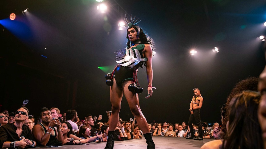 A vogue performer wearing a black-and-green outfit with a prominent silver fern poses on the runway at Sissy Ball 2019.