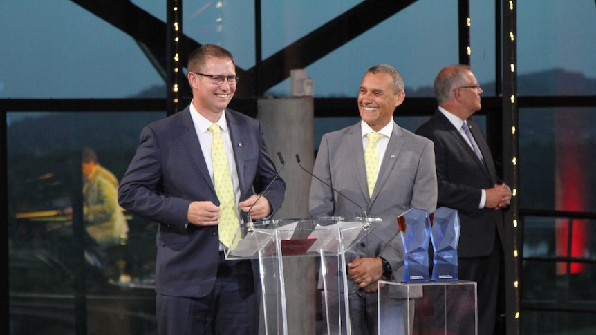 Two men in suits, Craig Challen and Richard Harris, stand at a podium in front of Scott Morrison. Two awards sit in front