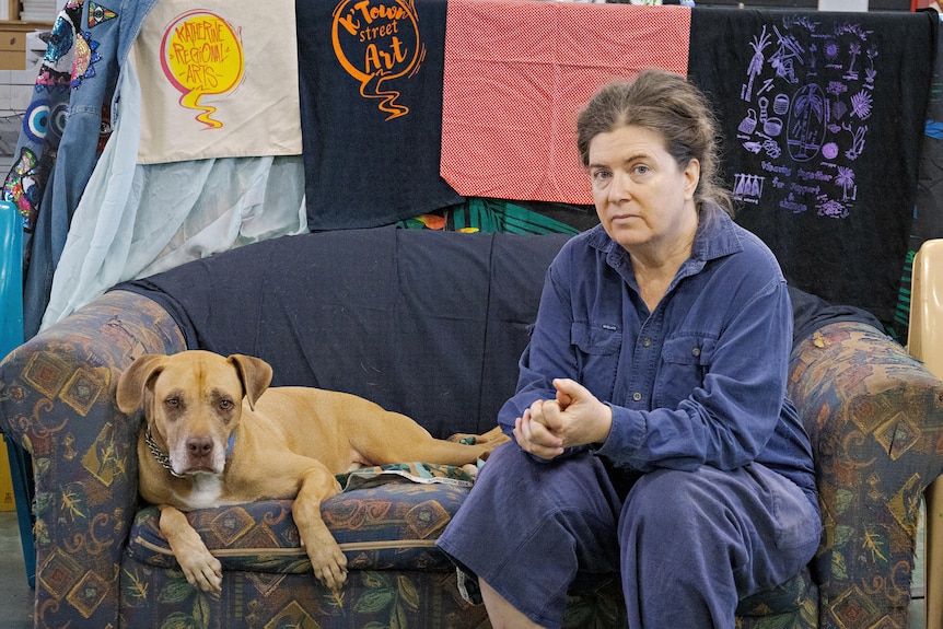 A woman in a navy blue jumpsuit sits on a couch next to a dog