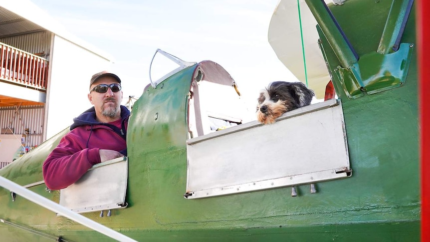 A small grey and white dog sits in the front cockpit of a green aircraft with a man in the cockpit behind him.