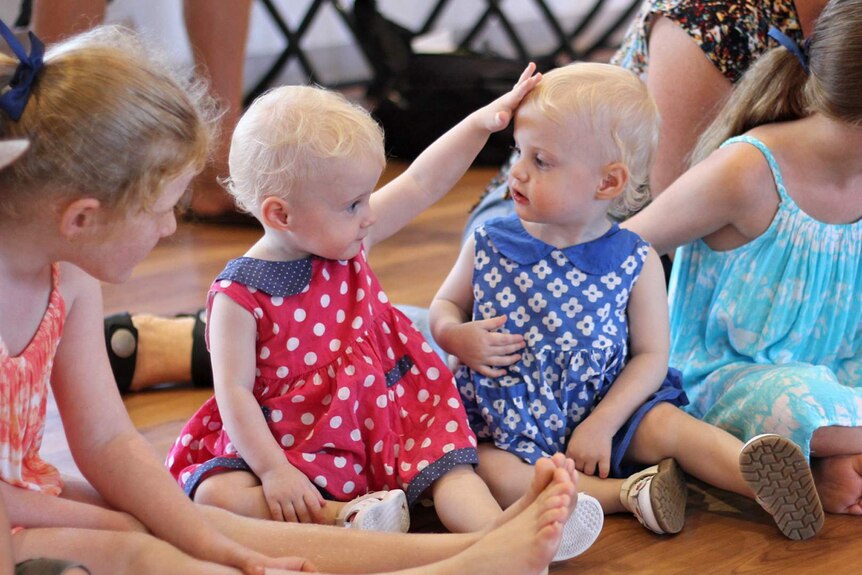 A blonde baby touches the head of her twin sister